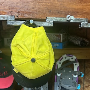 May include: A collection of hats, including a bright yellow cap, hang from metal hooks attached to a wooden beam. Other hats with black and red designs are also visible. A pair of snowshoes and antlers are also in the image.