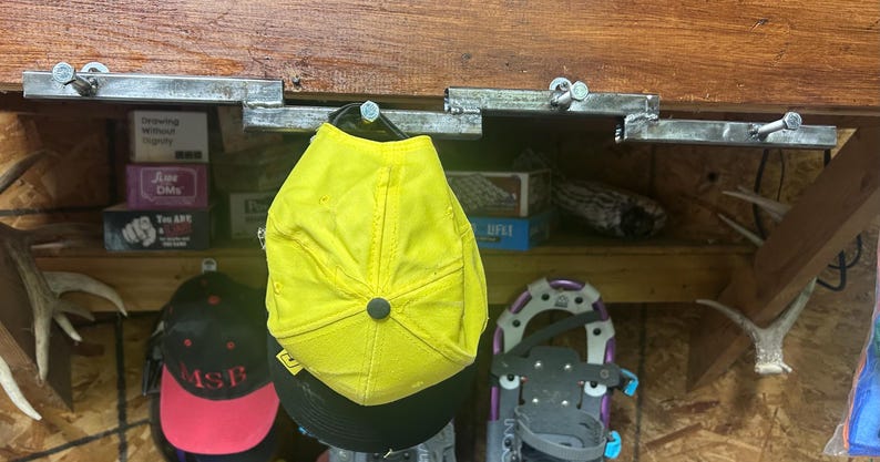 May include: A collection of hats, including a bright yellow cap, hang from metal hooks attached to a wooden beam. Other hats with black and red designs are also visible. A pair of snowshoes and antlers are also in the image.