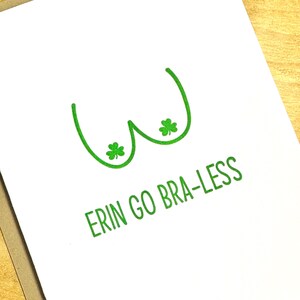 May include: White greeting card with a green line drawing of a bra and two shamrocks. The text "ERIN GO BRA-LESS" is printed in green below the drawing. The card is on a wooden surface.
