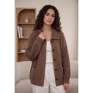 May include: A brown, long-sleeved, ribbed knit cardigan featuring a collared neckline, a button-down front with five dark buttons, and two front patch pockets. The garment is worn over a plain white top and light-coloured trousers.