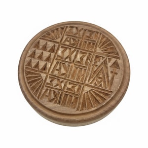 May include: A round, wooden butter mold with a detailed geometric design carved into its surface. The design includes triangles, squares, and radiating lines. The mold is light brown and appears to be handmade.