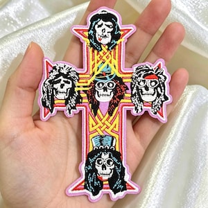 May include: A pink cross-shaped patch featuring five skull illustrations with long hair and various accessories. The patch has a pink border and yellow and red accents, with a Celtic knot design in the center.