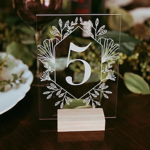 May include: Clear acrylic table number 5, with a white floral design. The number is displayed on a clear acrylic sheet, supported by a small, natural wood base. Ideal for weddings or events, adding a touch of elegance.