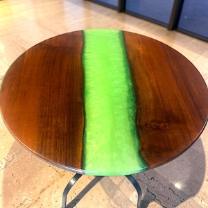 May include: A round table featuring a polished dark brown wood surface divided by a wide, vibrant neon green resin river inlay. The table is supported by four curved black metal legs that extend outward from the center base to the floor.