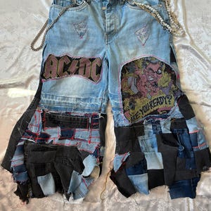 Pode incluir: Calças de ganga azul-claro com design de retalhos, bordas desfiadas, cinto de corrente prateada e emblemas AC/DC. A frente apresenta um gráfico de um diabo com o texto ARE YOU READY? e vários quadrados de tecido de ganga azul-escura e preta.