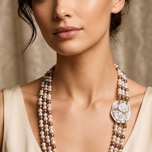 Peut inclure: Un collier multi-rangs composé de trois rangées de perles alternant entre le crème, le brun clair et le brun foncé. Les brins sont reliés sur un côté par un pendentif camée floral blanc et circulaire, sculpté de détails de pétales.