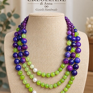 Peut inclure: Un collier à plusieurs rangs composé de perles facettées violettes et bleues, alternées avec des perles lisses vert citron et trois perles blanches. Le collier est présenté sur un mannequin beige avec le texte CREOLLANE di Anna Gioielli Handmade.