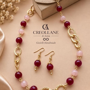 May include: A necklace and earring set featuring faceted deep red and pale pink beads interspersed with textured gold-tone circular links. The necklace includes a heart-shaped clasp. Text reads: CREOLLANE di Anna Gioielli Handmade.