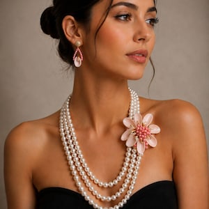 Pode incluir: Uma mulher usando um colar de pérolas de três fios com um broche de flor rosa. Brincos rosa e dourados combinando completam o conjunto. A mulher está usando um top preto sem alças.