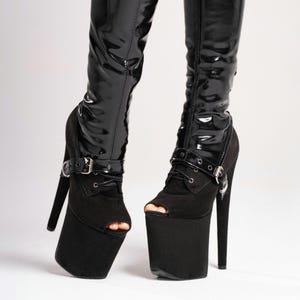 Könnte beinhalten: Schwarze Plateau-Stiefel mit offenen Zehen. Die Stiefel haben einen hohen Stiletto-Absatz und eine dicke Plateausohle. Der obere Teil des Stiefels besteht aus glänzendem schwarzem Material, und der Knöchelbereich hat einen Schnallenverschluss. Modisches Design.