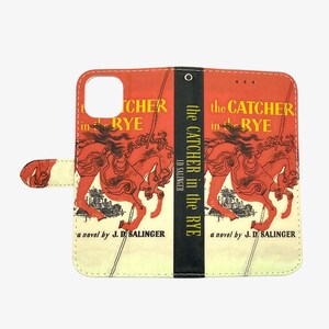 Pu&ograve; includere: Una custodia per telefono bianca e gialla con un'illustrazione rossa e nera di un cavallo di carosello. Il testo "the Catcher in the Rye" &egrave; stampato sulla custodia. Un romanzo di J.D. Salinger.