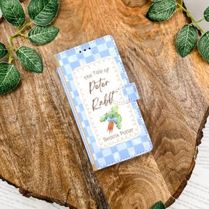 May include: A blue and white checkered phone case featuring the title "The Tale of Peter Rabbit" with an illustration of a carrot and Beatrix Potter's name. The case has a magnetic closure and is set on a wooden surface.