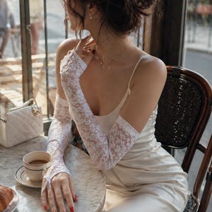 May include: A woman wearing a cream-colored slip dress and white lace gloves sits at a marble table in a cafe. A white purse, a cup of coffee, and a croissant are on the table. The woman is wearing a delicate necklace.