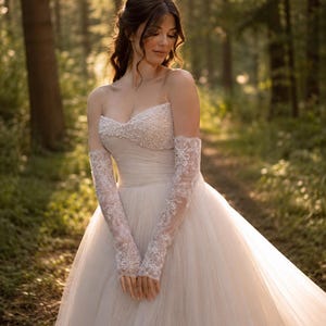 Könnte beinhalten: Ein elfenbeinfarbenes Brautkleid ohne Träger mit einem bestickten Oberteil und einem fließenden Tüllrock. Das Kleid hat zarte Spitzenärmel. Die Szene spielt in einem sonnigen Wald und schafft eine romantische und ätherische Atmosphäre.