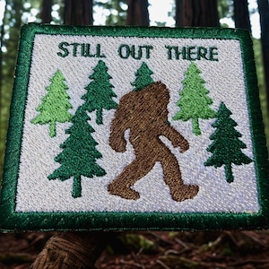 May include: A square embroidered patch with a white background and a dark green border. The patch features a brown silhouette of a creature walking among several green pine trees. The text "STILL OUT THERE" is embroidered in dark green letters.