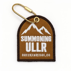 May include: A brown, arch-shaped embroidered patch keychain with a gold-toned metal carabiner. The patch features white embroidered mountain peaks above the text "SUMMONING ULLR" and "BRECKENRIDGE, CO" in a bold, sans-serif font with a dark brown border.