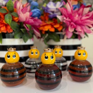 May include: A collection of decorative bee-shaped bottles. Each bottle has a yellow head with a crown and a smiling face. The bodies are brown with black stripes. The background features colorful flowers in black and white striped vases.