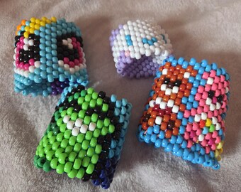 Può includere: Quattro braccialetti di perline colorate. Uno è azzurro con una faccia da cartone animato, un altro è bianco e viola, un terzo è verde e nero, e l'ultimo è arancione, rosa e blu. Questi braccialetti fatti a mano sono perfetti per aggiungere colore.