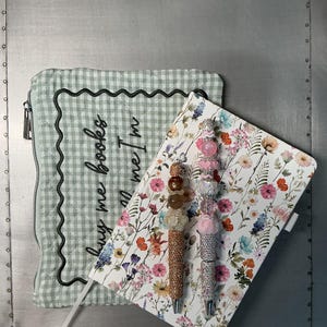 May include: A light green gingham zippered pouch with the embroidered text "buy me books and tell me I'm pretty", a floral notebook, and two beaded pens. One pen features brown and gold beads, while the other has pink and iridescent beads.