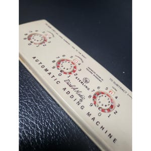 May include: An off-white Automatic Dial-A-Matic Adding Machine with three circular dials. The dials have red and white markings. The text "Automatic Adding Machine" is printed on the device.