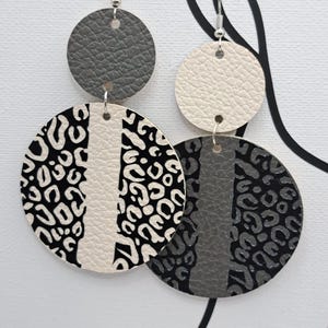 May include: A pair of dangling earrings with a layered design. The top circles are gray and off-white. The lower circles feature a black and white leopard print and a gray and black leopard print, with a vertical stripe of off-white and gray.