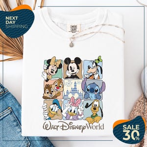 A white t-shirt with a graphic design featuring various Disney characters, including Minnie Mouse, Mickey Mouse, Goofy, Donald Duck, and Pluto, arranged in a grid pattern.