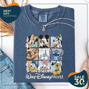 A t-shirt with a graphic of Disney characters, including Minnie Mouse, Mickey Mouse, Goofy, Donald Duck, and Pluto, arranged in a square pattern. The t-shirt is displayed on a white background, and there are jeans and a sweater visible in the background.