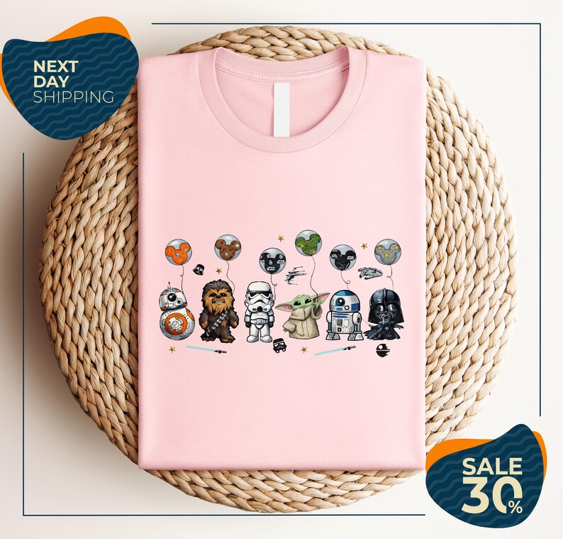 A pink t-shirt with a graphic design featuring various Star Wars characters, including Chewbacca, Stormtrooper, Darth Vader, and Baby Yoda, along with balloons and stars.