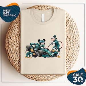 May include: A beige t-shirt featuring a graphic of Mickey Mouse in a green Formula 1 race car, with Goofy, Pluto, and Donald Duck dressed in matching green pit crew uniforms. The car displays the number 14 and Aston Martin branding.
