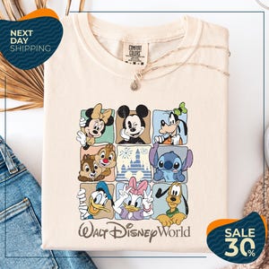 A white t-shirt with a graphic design of Disney characters, including Minnie Mouse, Mickey Mouse, Goofy, Donald Duck, and Pluto, arranged in a grid pattern. The t-shirt is displayed on a white background, and there are jeans visible in the background.