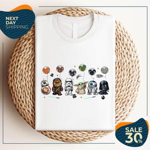 A white t-shirt with a graphic design featuring various Star Wars characters, including Chewbacca, Stormtrooper, Darth Vader, and Baby Yoda, along with balloons and stars.