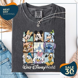 A gray t-shirt with a graphic of Disney characters, including Minnie Mouse, Mickey Mouse, Goofy, Donald Duck, and Pluto, arranged in a square pattern. The t-shirt is displayed on a white background, and there are jeans and a necklace visible in the image.