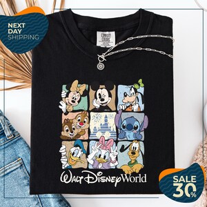 a black t-shirt with a graphic of Disney characters, including Minnie Mouse, Mickey Mouse, Goofy, Donald Duck, and Pluto, arranged in a square pattern. The t-shirt is displayed on a white background, and there are jeans and a necklace visible in the foreground.
