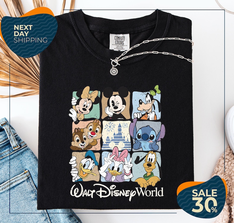 a black t-shirt with a graphic of Disney characters, including Minnie Mouse, Mickey Mouse, Goofy, Donald Duck, and Pluto, arranged in a square pattern. The t-shirt is displayed on a white background, and there are jeans and a necklace visible in the foreground.