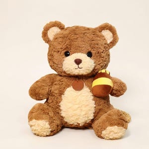 Könnte beinhalten: Ein braunes Teddybär-Plüschtier mit hellbraunem Bauch und Innenohren. Der Bär hat eine kleine braune Nase und schwarze Augen. Er hält einen braunen und gelben Honigtopf. Ein kleines braunes Etikett ist an der Brust des Bären befestigt.