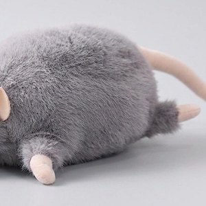 Könnte beinhalten: Ein graues Plüschtier in Form einer Ratte mit einem langen, rosa Schwanz. Das Spielzeug hat kleine rosa Ohren und Pfoten. Die Ratte liegt auf einer hellgrauen Oberfläche. Das Spielzeug besteht aus weichem, flauschigem Material.