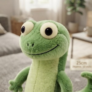 May include: A plush lizard toy with soft green fur, large cream-colored eyes with black pupils, and a stitched smile. The toy features a light green belly and a textured surface. A small sign in the background displays the text 25cm.
