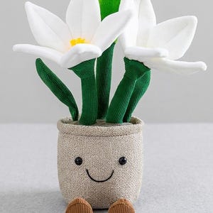 May include: A plush, smiling flower pot with white flowers, green stems, and brown feet. The pot is beige with a stitched face. The flowers have yellow centers. A whimsical, decorative item.