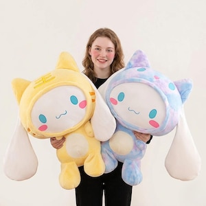 Puede incluir: Dos peluches, uno amarillo y otro azul, son mostrados. El juguete amarillo tiene una capucha con forma de gato con un diseño de tigre, el azul un patrón tie-dye. Ambos tienen orejas grandes, ojos redondos y mejillas rosas.