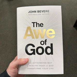 Puede incluir: Un libro titulado "The Awe of God" de John Bevere. La portada blanca presenta texto dorado y líneas radiantes. El título está en letras doradas grandes, con el nombre del autor en la parte superior. El subtítulo también es visible.