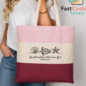 May include: A color-blocked tote bag featuring horizontal stripes in light pink, cream, and dark maroon. The center cream stripe displays a black line-art illustration of a seashell, a sea turtle, and a starfish, with the text Be still and know that I am God PSALM 46:10.