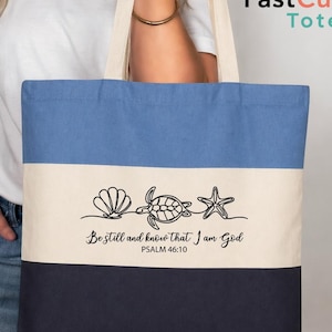 May include: A canvas tote bag with a blue, white, and navy color block design. The bag features a seashell, turtle, and starfish graphic with the text "Be still and know that I am God" and Psalm 46:10.