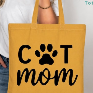 May include: Mustard yellow tote bag with black text and a paw print. The text reads "CAT mom". The bag has two handles and is made of a sturdy fabric. The bag is being held by a person.