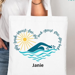 May include: White tote bag featuring a heart-shaped design with a swimmer illustration, sun, and water waves. The words "breast, free, fly, back" are written around the heart. The name "Janie" is printed below the design. The bag has long handles.