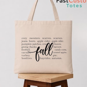 May include: A beige canvas tote bag featuring black printed text that lists various autumn-themed words including cozy sweaters, scarves, jeans, boots, apple cider, pumpkin patches, leaves, giving thanks, harvest, football, candy corn, turkey, and bonfires.