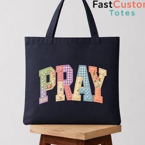May include: A navy blue tote bag featuring the word PRAY in large, quilted, multi-colored fabric letters. Each letter displays a different pattern, including floral prints, gingham checks, and polka dots in pastel shades of green, pink, blue, and yellow.
