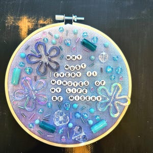 May include: A round embroidery hoop with a light blue fabric background. The text "WHY MUST EVERY 00 MINUTES OF MY LIFE BE MISERY" is spelled out with white letter beads. The design includes blue and clear beads and flower-shaped embellishments.