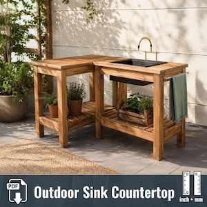 May include: An L-shaped outdoor wooden workstation featuring a black sink basin, a gold-toned tap, and a side towel bar with a green towel. The unit includes lower slatted shelves holding potted plants and a wooden crate with greenery.