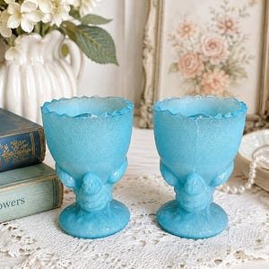 May include: A pair of light blue glass egg cups featuring a textured surface and a scalloped rim. Each cup is supported by a base molded into the shape of a small bird, with the bird's head and wings visible on the stem of the vessel.
