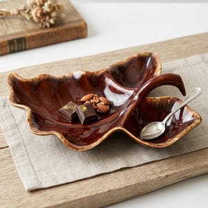 May include: A brown ceramic leaf-shaped serving dish with a handle, holding chocolate squares, almonds, and a small silver spoon. The dish has a glossy finish and a textured rim, perfect for serving snacks.